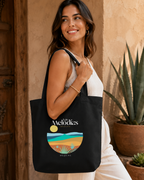A woman wearing a white sleeveless top and white pants carries a black canvas tote bag with a colorful landscape graphic and the text 'Melodies by Mezcal'. The setting features a warm, rustic beige stucco wall, a wooden door, and a potted agave plant.