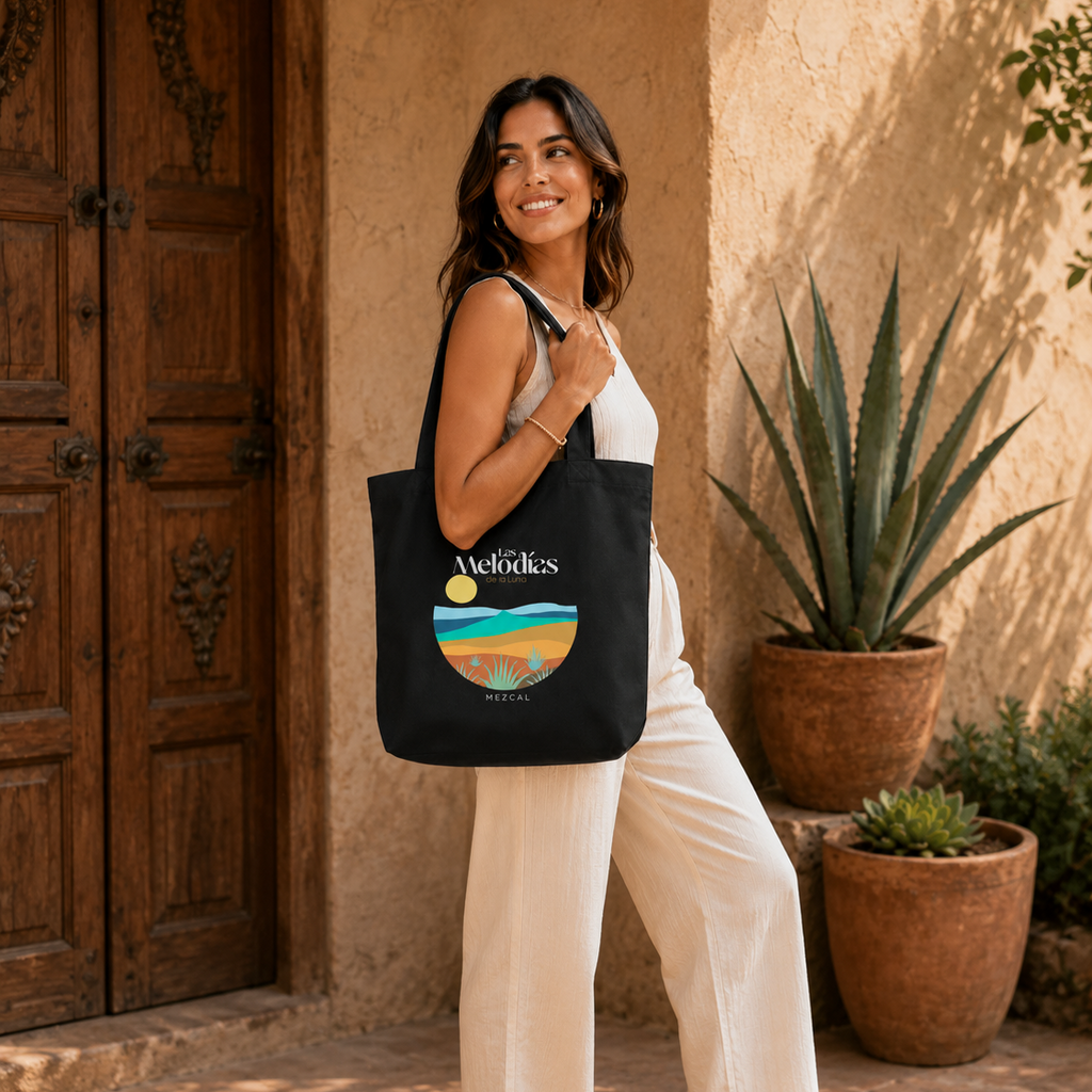 A woman stands in a sunlit courtyard wearing a white sleeveless jumpsuit and sandals, carrying a black Las Melodias tote bag with a colorful retro beach graphic and the word 'Vacation' printed on it. The background features a warm stucco wall, a large wooden door, and potted agaves.