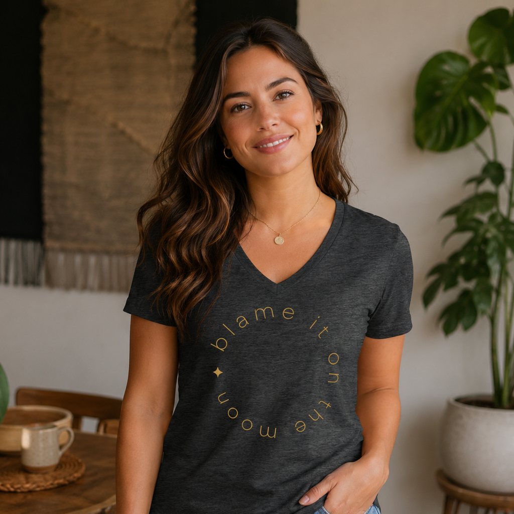 A woman wearing a dark charcoal gray V-neck t-shirt with a gold graphic that reads 'blame it on the moon' in a circular arrangement, paired with light blue jeans, standing in a cozy indoor setting with plants and wooden furniture.