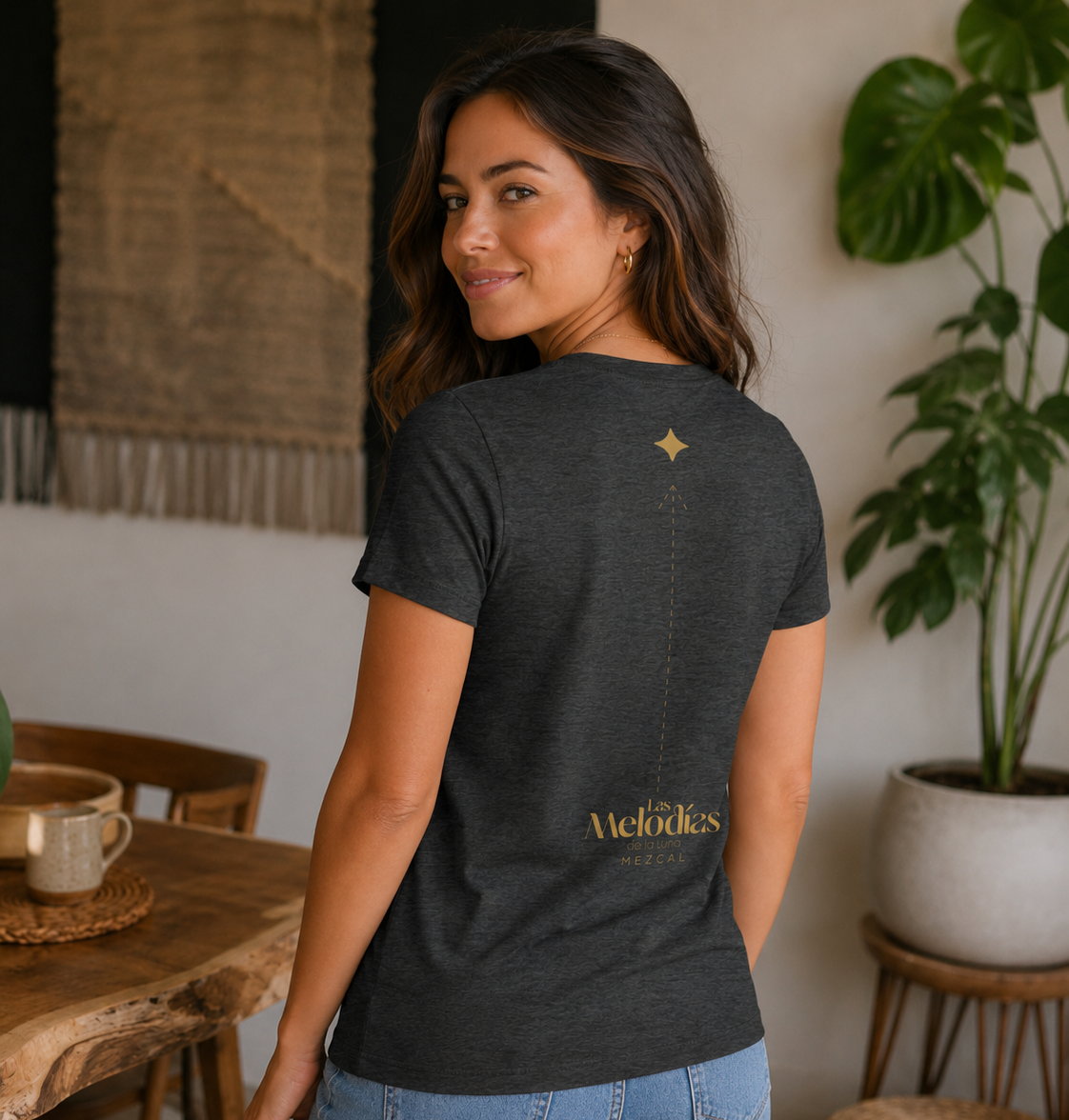 A woman wearing a dark heather gray t-shirt with a gold 'Melodies' logo on the back, paired with light blue jeans, standing in a cozy, modern room with plants and wooden furniture.